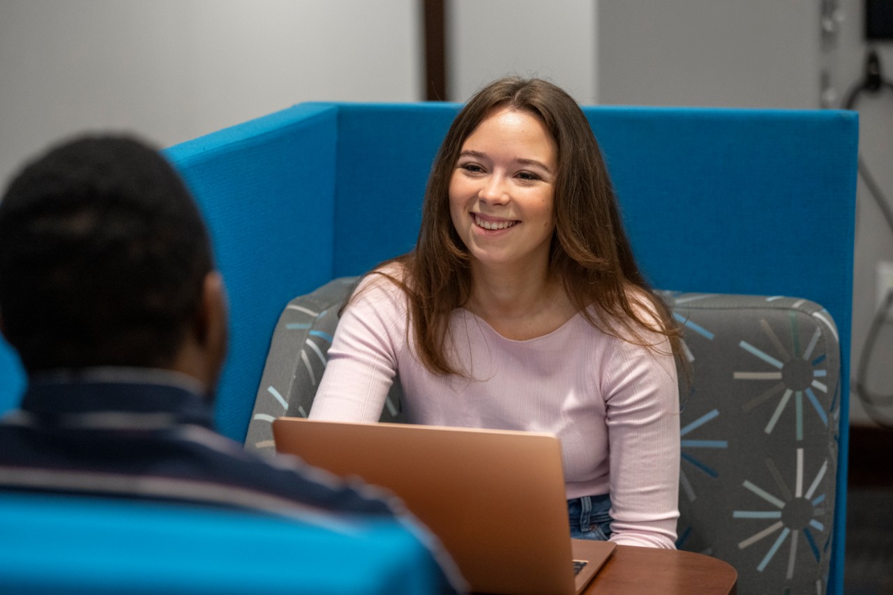 Person sitting and looking forward and talking to another student with a laptop in front of them.